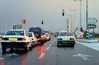 בתמונה נראה כביש רטוב בשעת דמדומים או במזג אוויר סגרירי, היוצרים תנאי ראות לקויה. מספר כלי רכב עומדים בטור לפני צומת מרומזר, והרמזורים דולקים באור אדום.