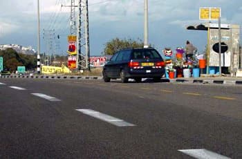בצד ימין של הכביש נראה מפרץ המיועד לתחנת אוטובוס. בתוך המפרץ חונה רכב פרטי מסוג סטיישן, ולידו עומד אדם עם דוכן למכירת פרחים. אבני השפה של המדרכה צבועות בשחור-לבן.