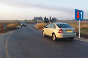 כביש דו-סטרי עם רכב קרוב ורכב רחוק המגיע מהכיוון הנגדי. בצד הדרך מוצב תמרור מלבני כחול עם שני חיצים אנכיים: חץ אדום הפונה כלפי מעלה וחץ לבן הפונה כלפי מטה.