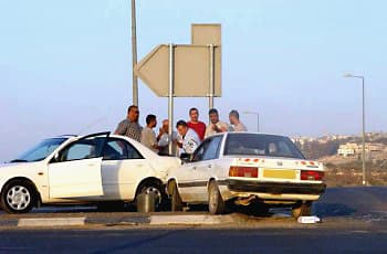 תמונה של תאונת דרכים בין שתי מכוניות פרטיות העומדות בשול הדרך. ליד כלי הרכב הפגועים עומדת קבוצת אנשים. ברקע נראה גב של תמרור הכוונה גדול בצורת חץ.