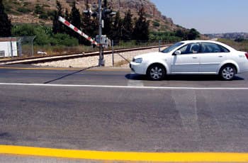 רכב לבן עצר לפני קו עצירה בכביש, מול מפגש מסילת ברזל. המחסום במפגש, בצבעי אדום-לבן, ירד וחוסם את הנתיב.