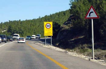בצד ימין של כביש בינעירוני מוצבים שני תמרורים. התמרור הקרוב הוא תמרור אזהרה משולש עם מסגרת אדומה, המציג סמל של עקומה חדה ימינה. מאחוריו ניצב תמרור מלבני צהוב, שבחלקו העליון עיגול עם המספר 90, ומתחתיו הכיתוב "סע בזהירות" ו-"REMINDER".