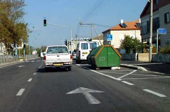 כביש עם חץ נסיעה ישר על האספלט ורמזור עם אור ירוק בהמשך. מימין חונים רכב מסחרי ומכולת אשפה גדולה, ולידם מוצב תמרור חניית נכה, שהוא עיגול כחול עם סמל כיסא גלגלים.
