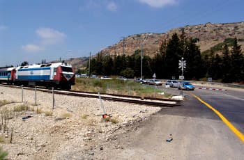 התמונה מציגה מפגש כביש עם מסילת ברזל. רכבת נוסעים נמצאת על המסילה, קרובה מאוד למפגש. מחסום בצבעי אדום-לבן ירד וחוסם את הכביש, ולידו ניצב תמרור בצורת X.