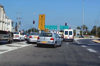 צומת מרומזר עם רמזורים אדומים. רכב פרטי כסוף עומד על שטח הפרדה מצויר בלבן, בין שני נתיבי נסיעה. מעל הנתיבים ישנו תמרור צהוב המורה על פניית פרסה מהנתיב השמאלי בלבד.