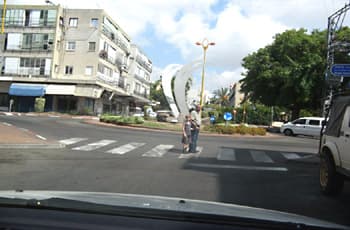 מנקודת מבט של נהג, רואים רכב המתקרב למעבר חצייה לפני כיכר תנועה. על מעבר החצייה, בנתיב הנסיעה של הרכב, עומדים מבוגר וילד. ליד הכיכר מוצב תמרור 303: עיגול כחול עם שלושה חצים לבנים המורים על תנועה מעגלית.