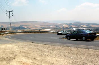 כביש דו-סטרי צר עם עקומה חדה שמאלה, ללא קו הפרדה מסומן בין הנתיבים. רכב כהה מתקרב לעקומה, ומולו, בתוך העקומה, נוסע רכב לבן. בצד החיצוני של הסיבוב מוצבים מספר תמרורי הכוונה (שברונים) המורים על כיוון הפנייה.