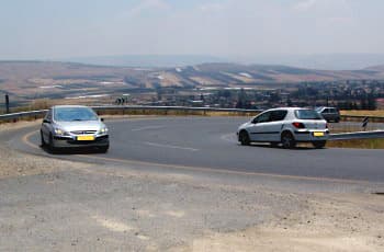 כביש דו-סטרי עם עקומה חדה שמאלה. שני כלי רכב נוסעים בעיקול, כל אחד בנתיבו, ושניהם צמודים לשפת הכביש הימנית.