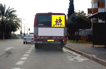 בתמונה נראה חלקו האחורי של אוטובוס שעוצר בצד ימין של הכביש. על האוטובוס מוצג שלט מרובע צהוב, עם צללית שחורה של ילדים הולכים והכיתוב "ילדים".