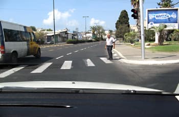 בתמונה נראה כביש עם מעבר חצייה, מנקודת מבט של נהג. הולך רגל עומד על מעבר החצייה, ליד מדרכה רגילה ללא סימון מיוחד. ליד המדרכה מוצב רמזור עם אור אדום דולק.