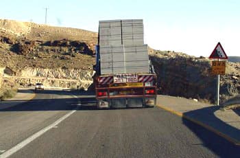 בתמונה נראית נסיעה מאחורי משאית כבדה בכביש מפותל עם פס הפרדה רצוף. בצד ימין של הדרך מוצב תמרור אזהרה משולש המזהיר מפני הידרדרות אבנים, ומתחתיו שלט צהוב המציין "הידרדרות אבנים למשך 1.5 ק"מ".