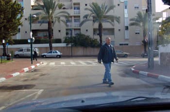 הולך רגל חוצה את הכביש, כשהוא נמצא במרכז נתיב הנסיעה של הרכב המצלם. מאחורי הולך הרגל, במרחק קצר, מסומן על הכביש מעבר חצייה.