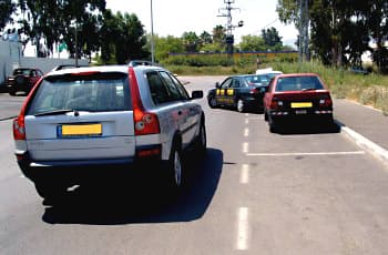 רכב כסוף נוסע בכביש או במגרש חנייה. לפניו, רכב שחור יוצא ממקום חנייה המסומן בקווים לבנים בצד ימין, וחלקו הקדמי כבר בולט לתוך נתיב הנסיעה של הרכב הכסוף.