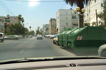 מבט מתוך רכב הנוסע ברחוב עירוני. בצד ימין של הדרך ניצבת שורה של מכולות אשפה ירוקות וגדולות, החוסמות את שדה הראייה למדרכה שמימין.