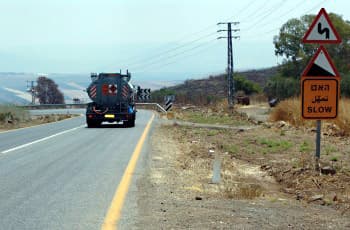 בכביש בינעירוני נראית מכלית שנוסעת בנתיב הימני, קרוב מאוד לקו הצהוב הרציף המסמן את שפת הכביש. מימין לכביש מוצבים שלושה תמרורים: למעלה תמרור אזהרה על פיתולים, מתחתיו תמרור אזהרה על ירידה מסוכנת, ולמטה תמרור כתום מלבני עם הכיתוב "האט, שוליים רכים" ו-"SLOW". השוליים לצד הכביש אינם סלולים.